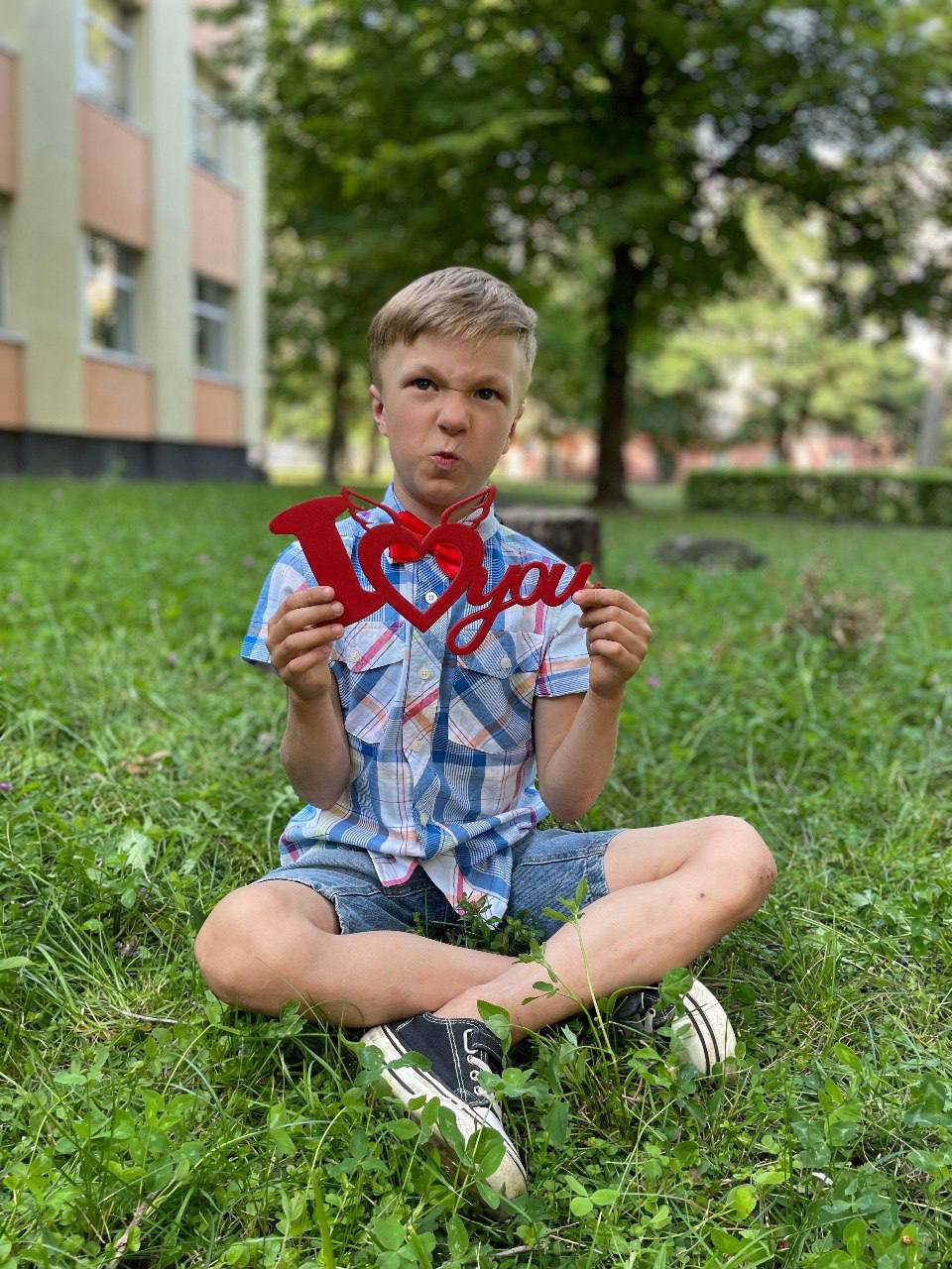 Хлопчик сидить на траві і кумедно кривляється з написом «I love you»